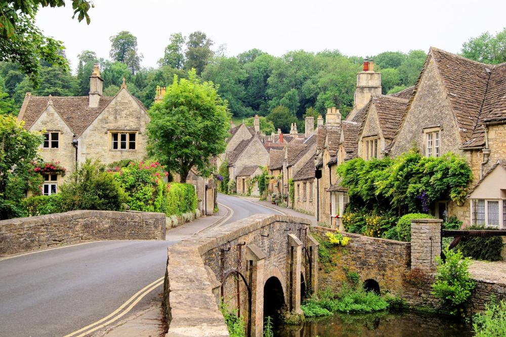 A picturesque bridge spans a river in a quaint Cotswolds village, surrounded by lush greenery and charming cottages