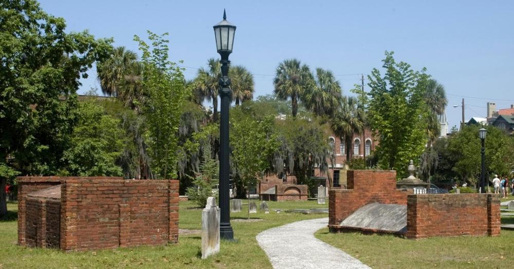 A brick wall and a light post stand in Colonial Park Cemetery, Savannah, surrounded by gravestones and greenery