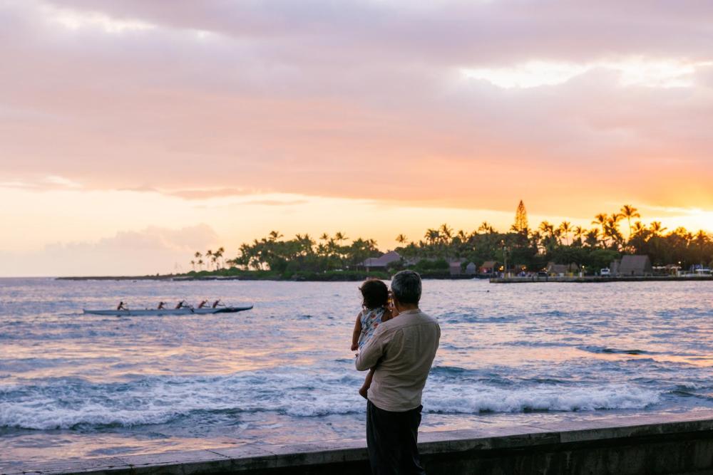 A man holds his child on a beach, gazing at the ocean during their Thanksgiving family trip in Hawaii