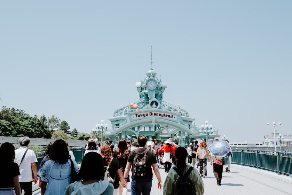 People walking on a bridge near entering Tokyo Disneyland, enjoying the vibrant atmosphere of the park. There's a blueish clocktower that has the sign "Tokyo Disneyland" in red letters.