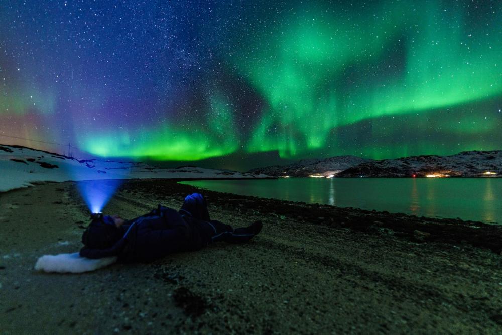 A person lying on the ground, gazing at the green-hued northern lights in Tromsø.