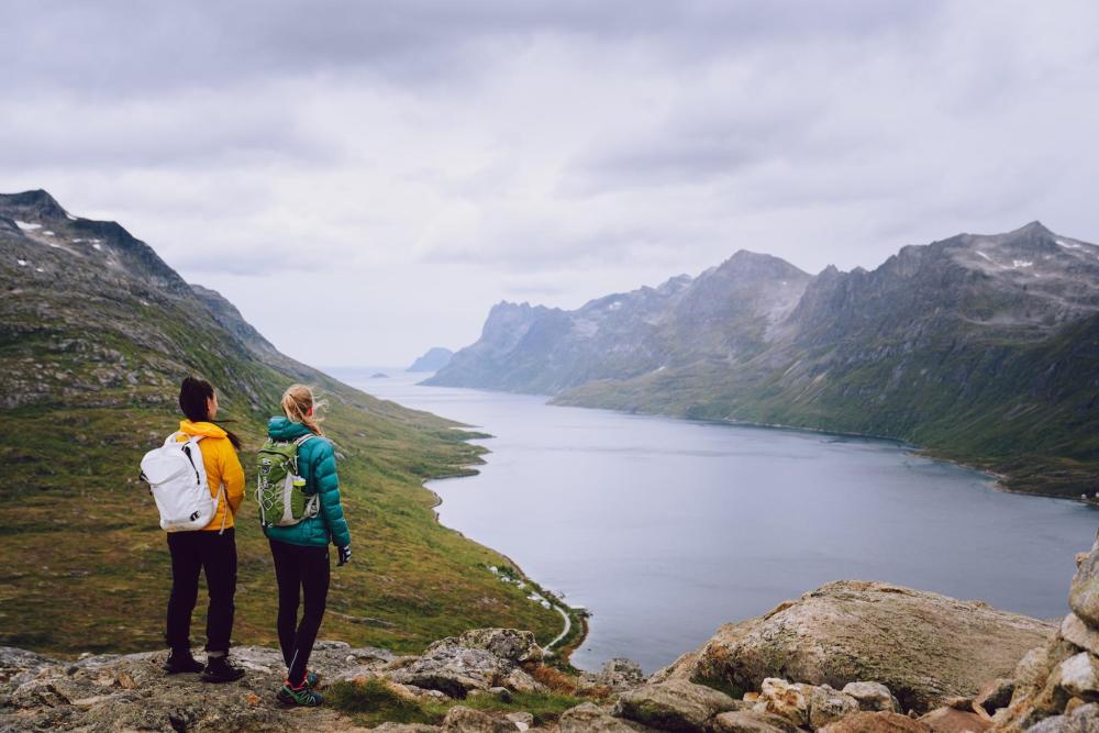 Two people stand on a mountain in Tromsø, overlooking a serene lake surrounded by mountains.