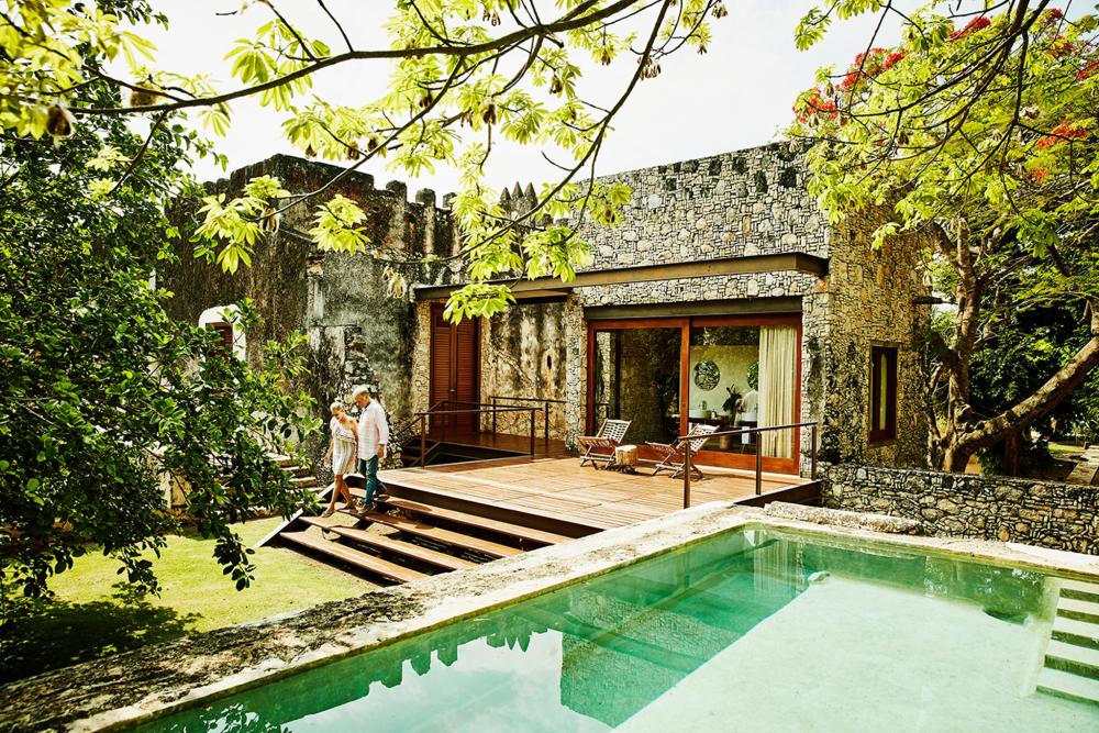 A man and woman stand together in front of a villa pool, enjoying a sunny day in a relaxed atmosphere.