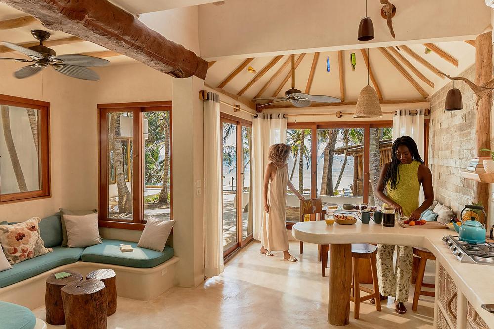 A woman is in the kitchen of a beach villa, featuring bright colors and a view of the ocean through the window.