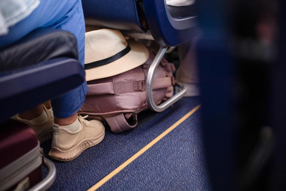 The airplane aisle seat, featuring a hat and bag stored under the seat in front.
