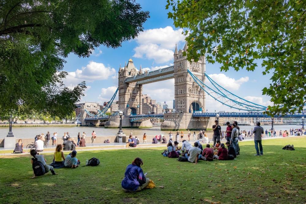 Tower Bridge stands tall on a sunny day in London, people sit on shaded grass near by.