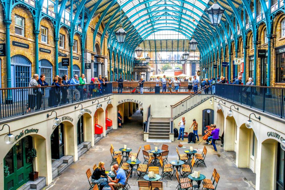 A wide scape shot of the ground and first floor of Covent Garden's Market. Visitors on the ground floor sit at tables eating and drinking, with those above admiring the views of the market.