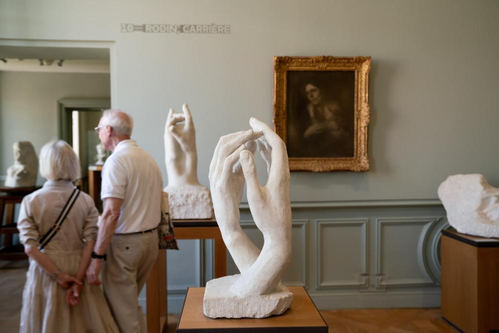 A hand-holding sculpture is featured in the foreground, with a couple admiring another piece at the Musée Rodin in the background.
