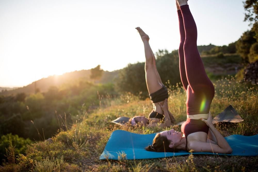 Two women practice a shoulder stand on a grassy hillside with a scenic view in the background.