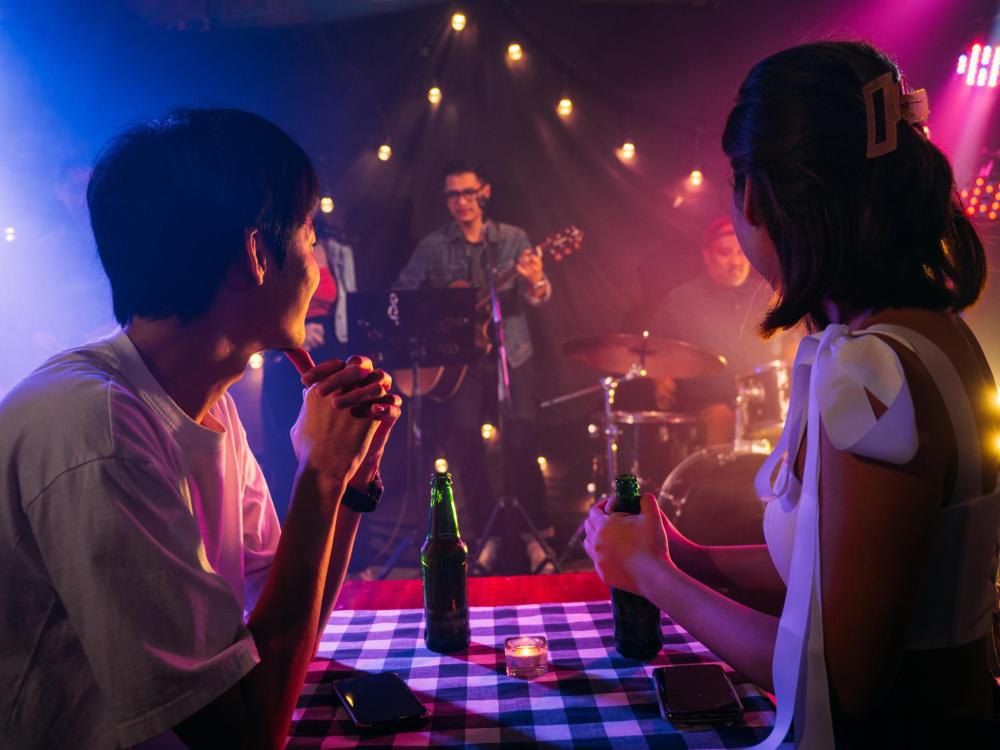 A couple sits at a table with drinks, watching a live band perform on a small stage in a cozy, dimly lit venue