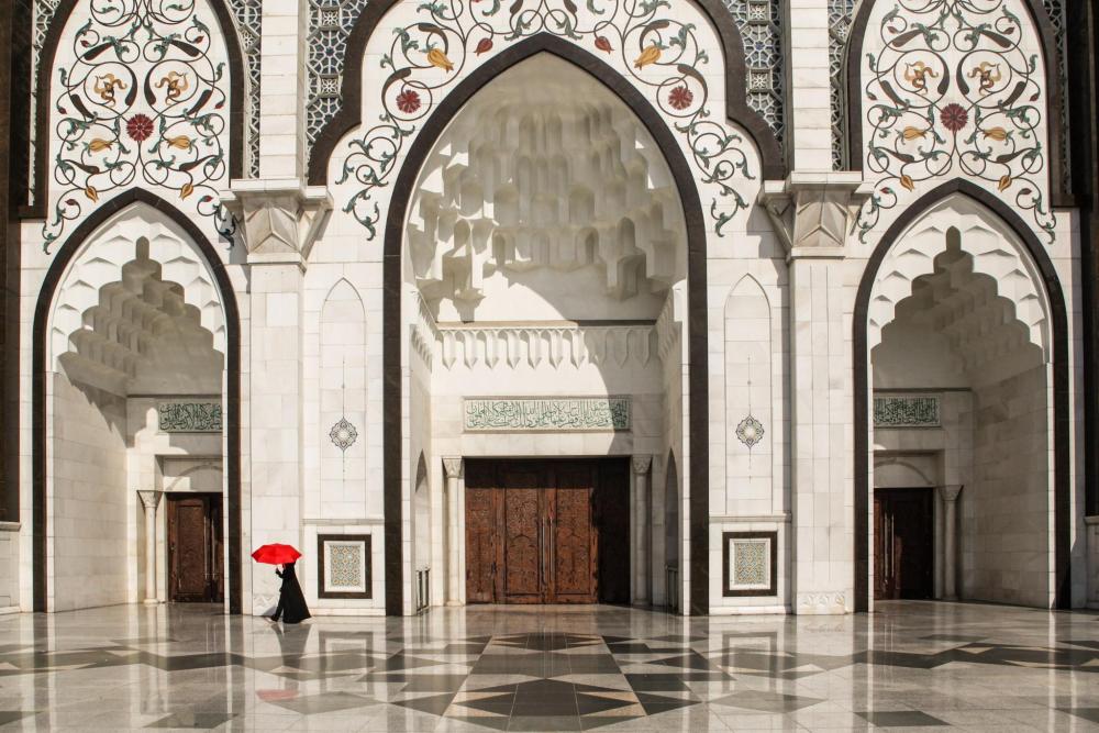 Grand mosque courtyard with ornate arches and geometric patterns, with a person walking through the open space.