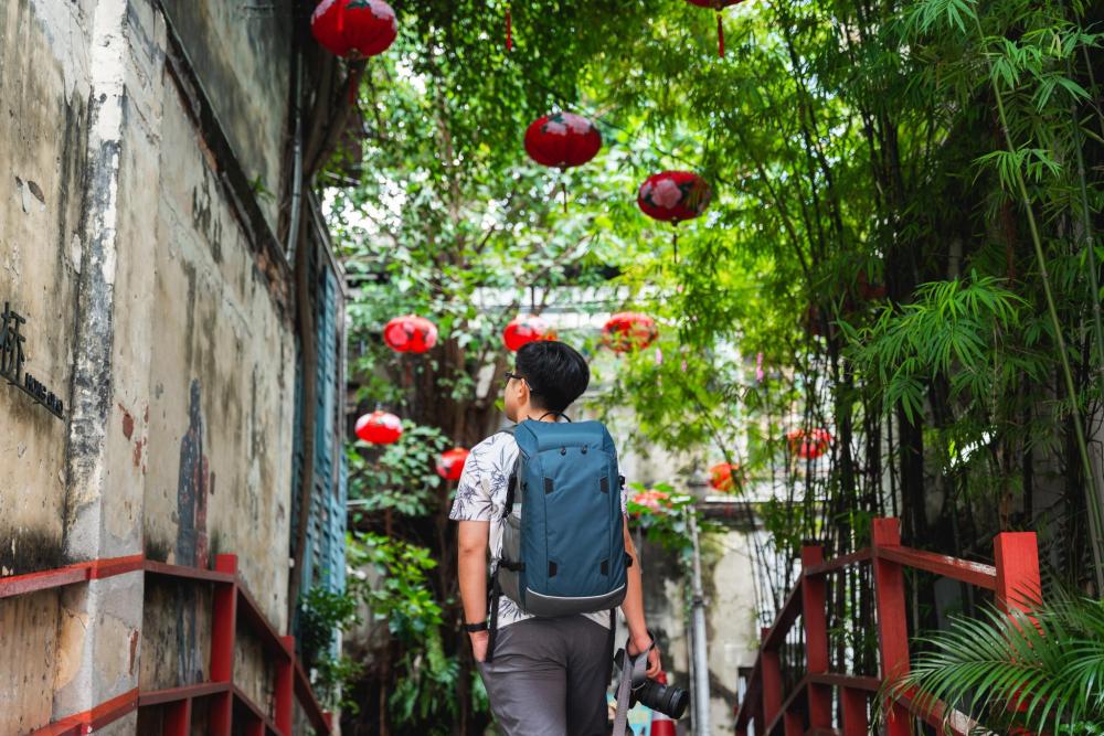 Traveller with backpack walking along a narrow street decorated with red lanterns and greenery in Kuala Lumpur.