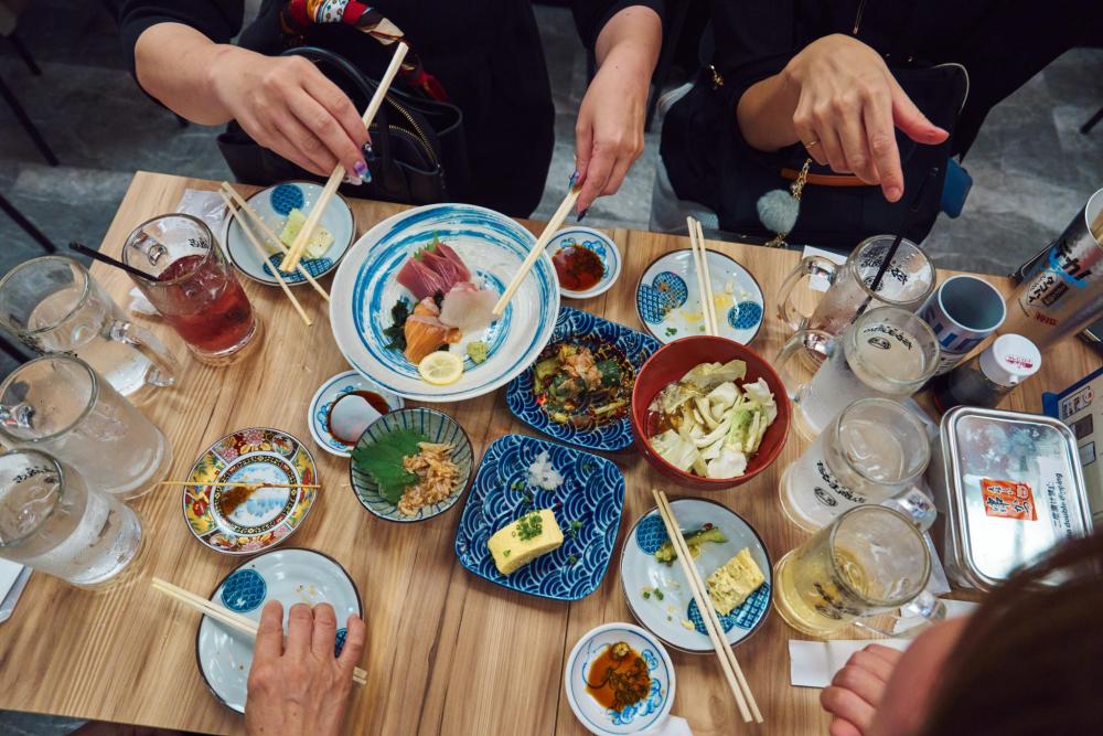 Group of people using chopsticks to share a Japanese meal with sashimi, small plates and drinks on a wooden table.