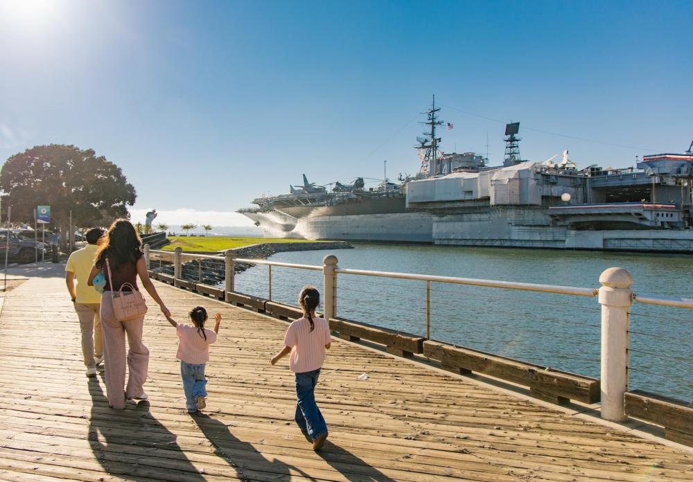 A family walks along a sunny wooden pier in San Diego, with a massive aircraft carrier docked in the background harbor.