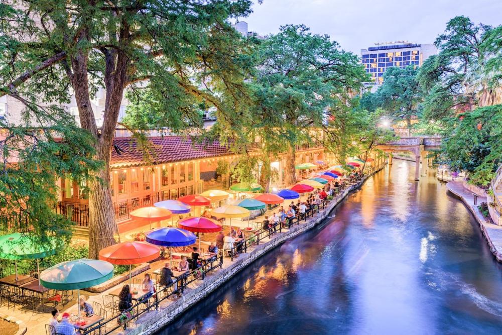 Vibrant San Antonio River Walk featuring colorful patio umbrellas, lush cypress trees, and diners along the water at dusk.