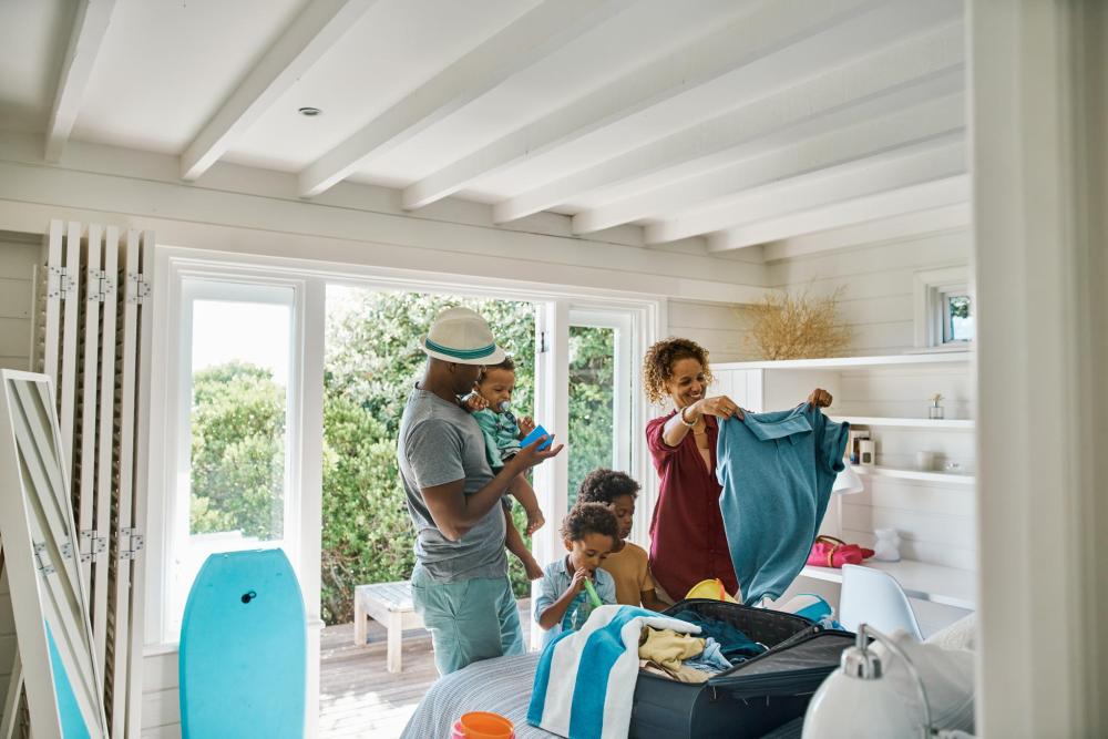 A family packs clothes in a suitcase together. The mother holds up a t-shirt as the father lifts up one of their three children.