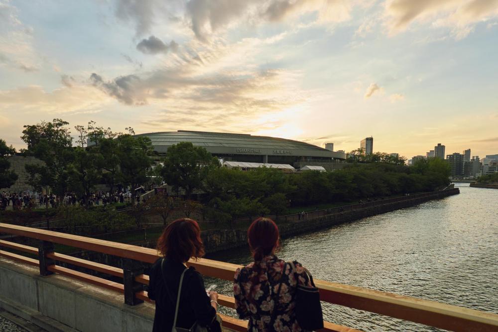 Two people looking over a river at sunset with Osaka-jō Hall and trees in the background