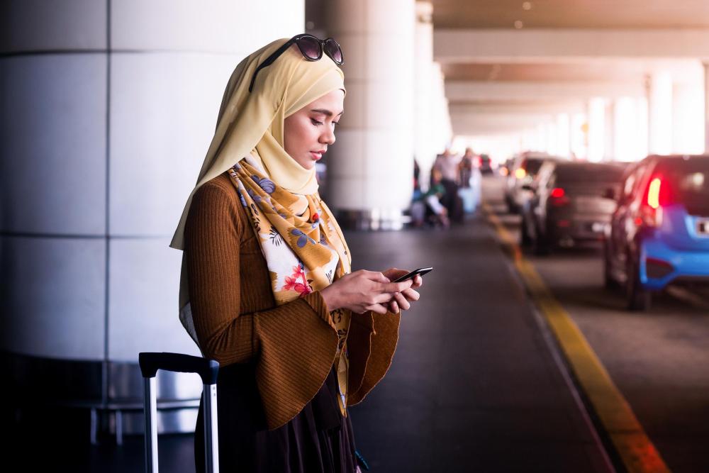 A woman wearing a headscarf stands beside her suitcase at an airport while checking her phone