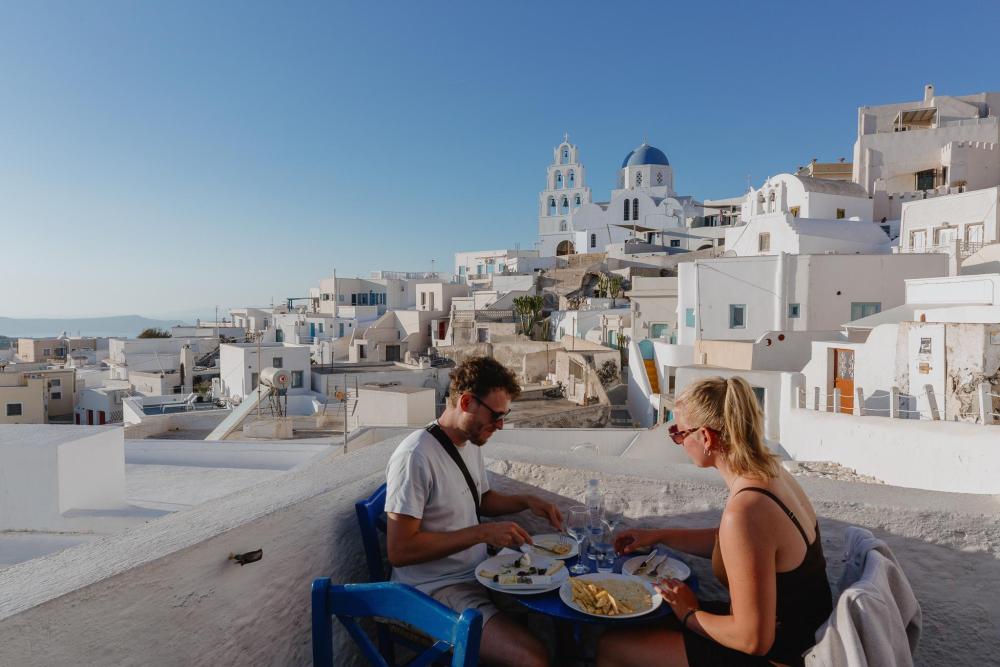 Couple dining on a terrace overlooking white buildings and a blue-domed church in Santorini