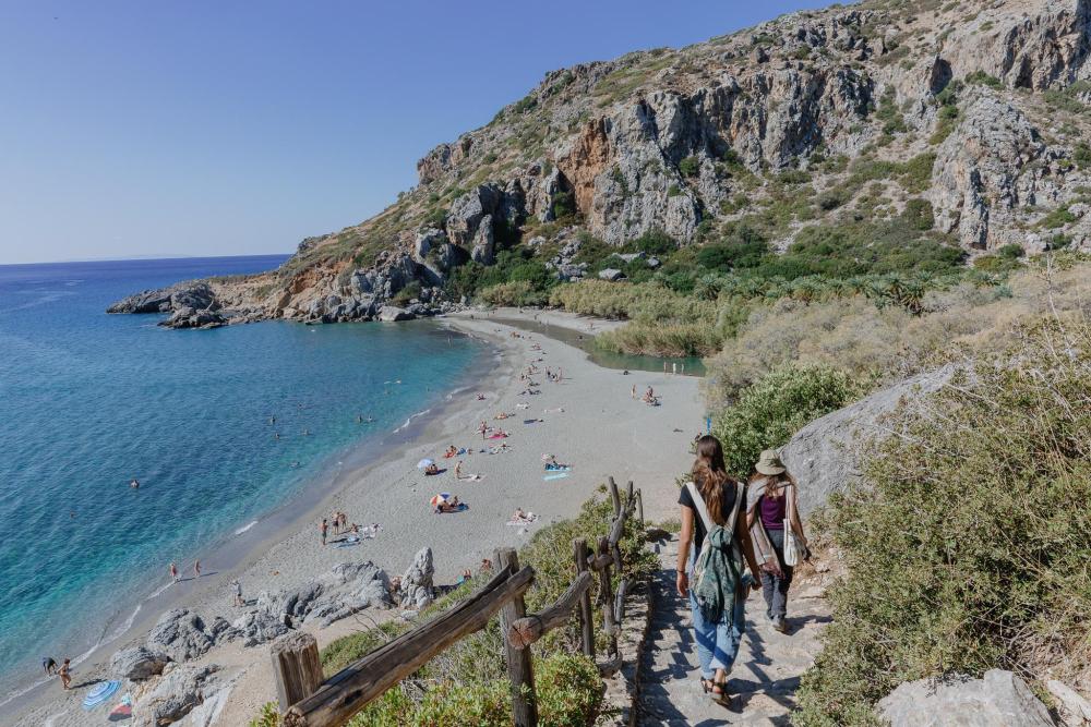 People walking down a path toward a quiet beach with turquoise water and rocky cliffs