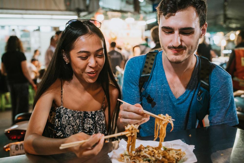 Two visitors sharing a street food dish at a busy Bangkok night market.