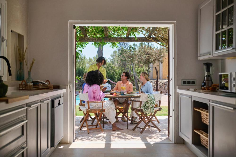 Four people share a meal on a sunny villa terrace, seen through open kitchen doors, with trees, a shaded pergola and relaxed holiday dining setting outside.