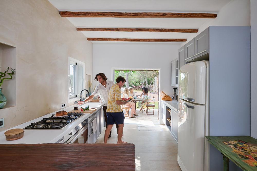 Two people prepare food in a bright villa kitchen while friends sit at a table outside on the sunny terrace, creating a relaxed holiday scene.