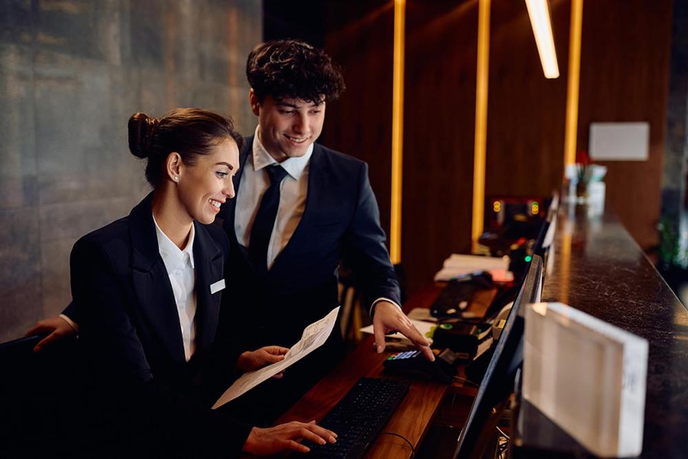 A man and woman in formal business attire are at a desk, collaborating on a project with documents and a laptop
