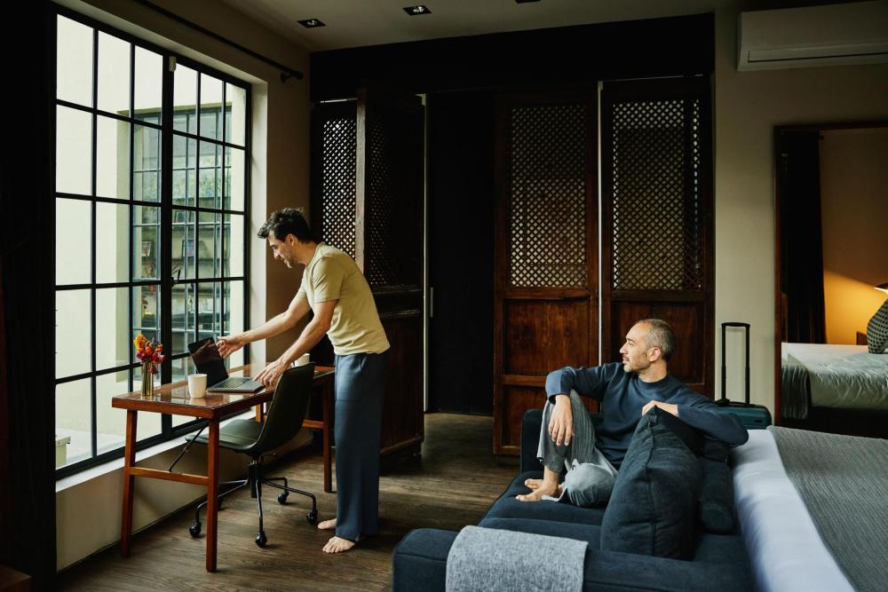 Two guests in a hotel room, one working at a desk by the window while the other relaxes on a couch next to the bed and suitcase.