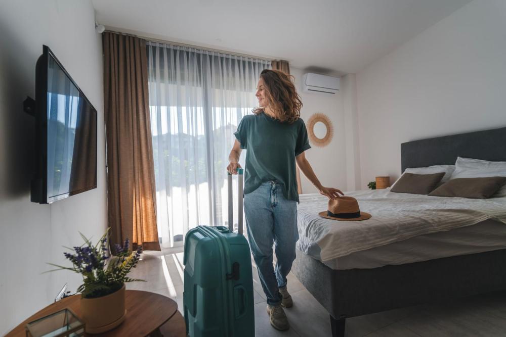 A woman stands with a suitcase in front of a neatly made bed, preparing for her journey.