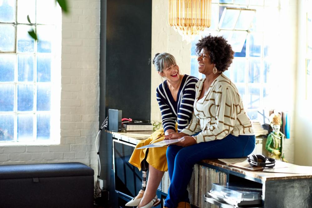 Two women laughing while sitting on a piece of furniture.