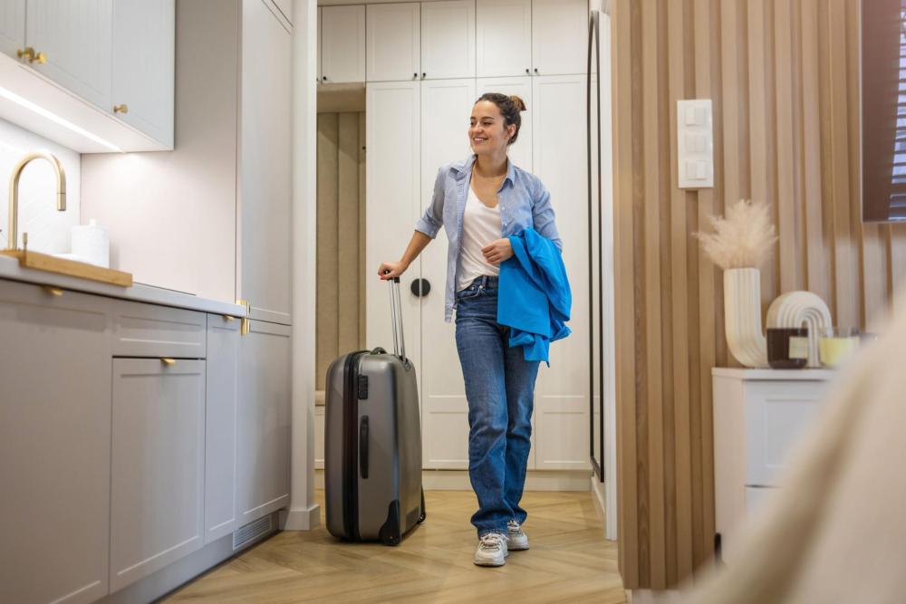 A smiling woman with a suitcase walks through a room with a kitchen.