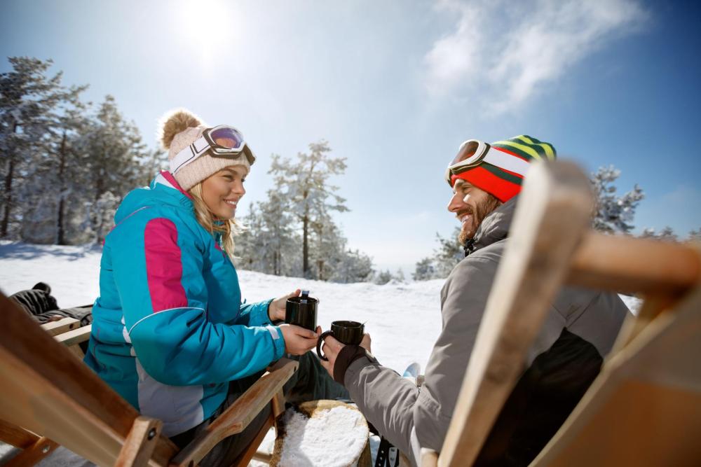A man and a woman sitting outside with hot drinks, smiling at each other while wearing ski gear.