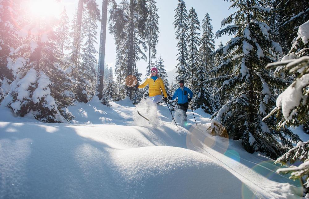 A man and a woman are walking with their skis in the snow in the middle of a forest, smiling