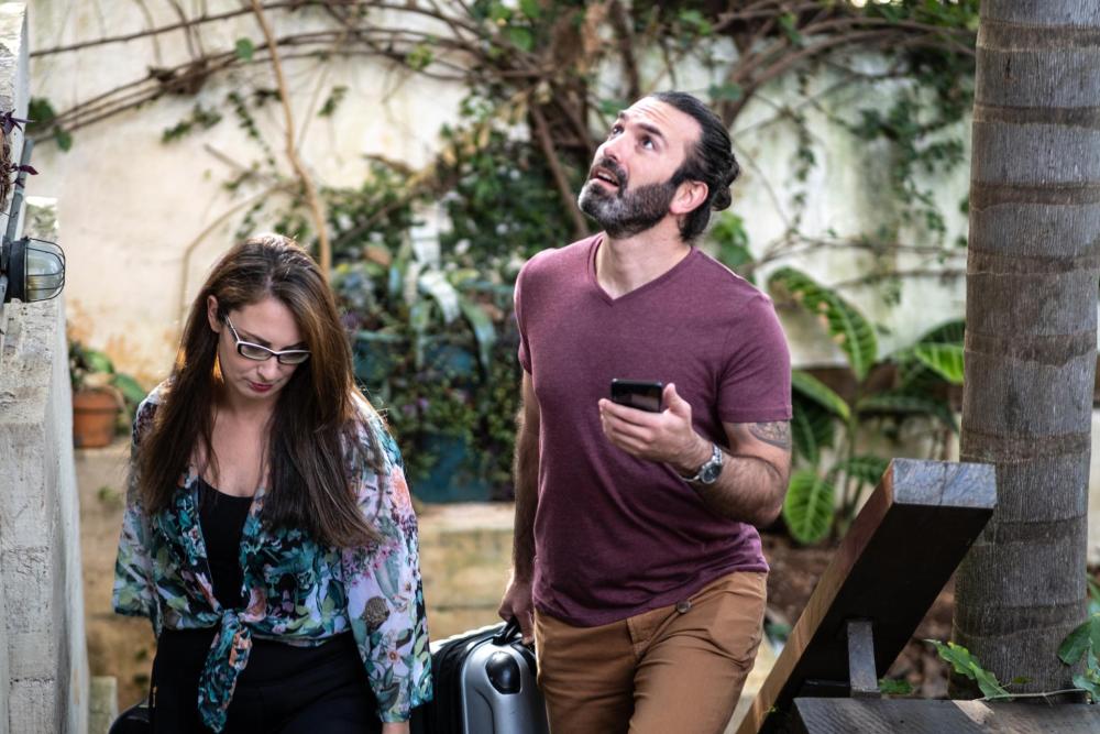 Two travellers walk up outdoor steps with suitcases, while one checks a phone in a leafy courtyard
