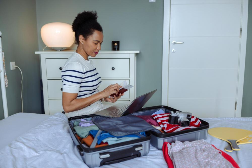 A person reviews their travel document and laptop while seated on a bed beside an open suitcase in a room