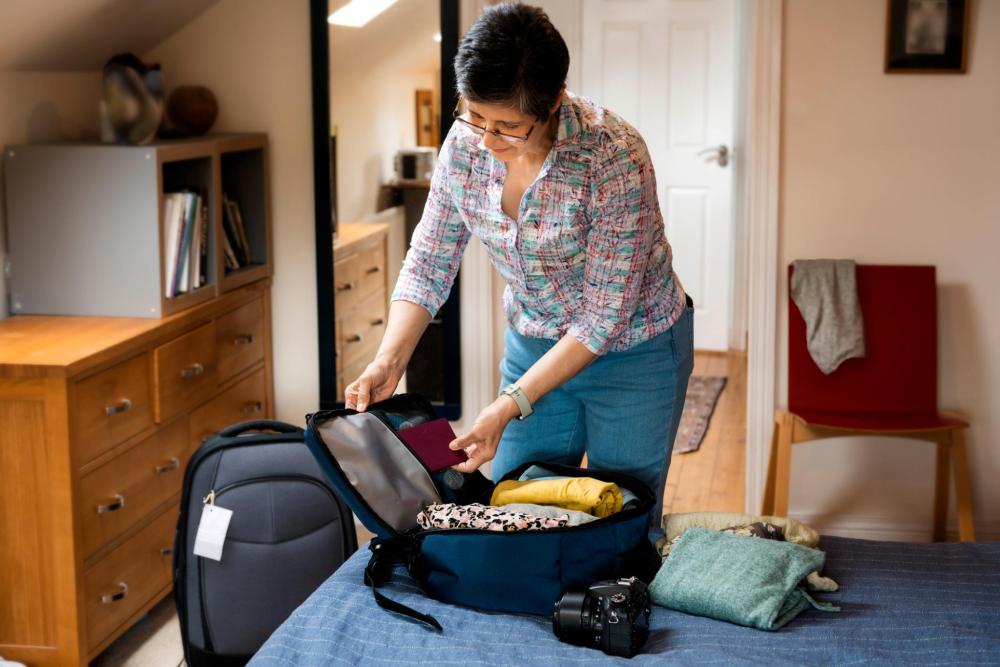 A person packs their luggage, placing their passport into a backpack next to an open suitcase in a bedroom