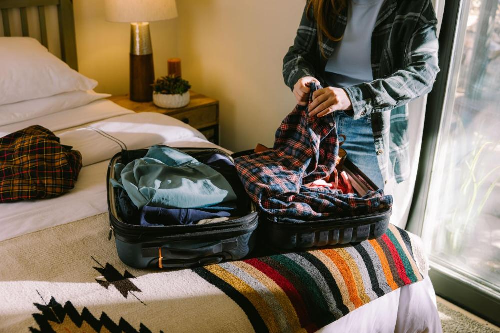 A person packing a plaid shirt into a suitcase on a bed in a hotel room.