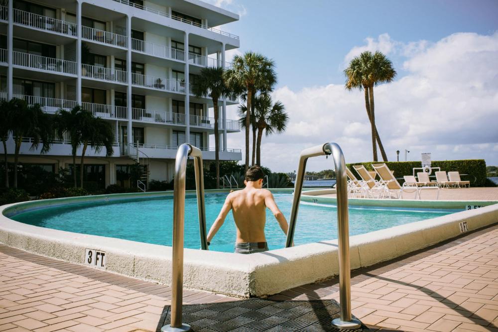 A teenager stepping into an outdoor pool beside a waterfront apartment building, with sun loungers and palm trees in the background.