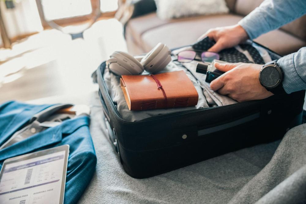 A person packing a suitcase with travel essentials like clothes, a notebook, and glasses.