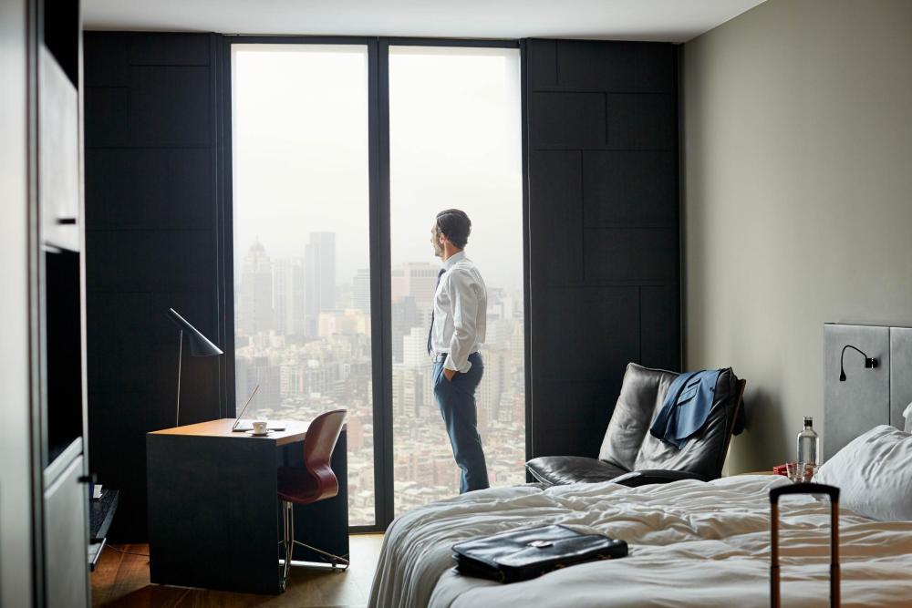 A business traveler looks out at an urban landscape from a hotel room.