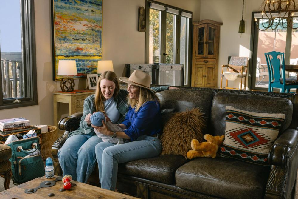 Two guests sit on a sofa in a cozy vacation rental, smiling while holding a baby, with travel bags and toys nearby