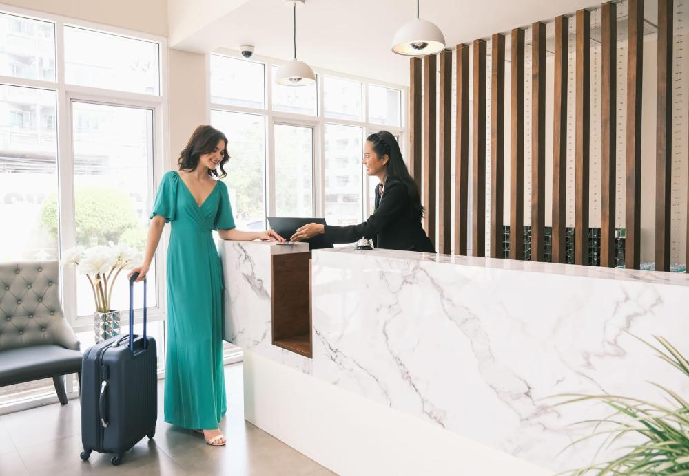A woman stands in a hotel lobby with her suitcase at the reception desk