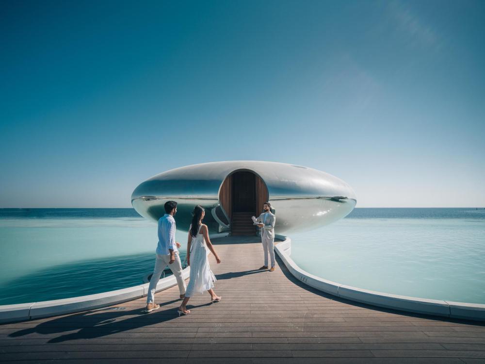 A couple walks along a sleek overwater walkway toward a futuristic pod-shaped villa at Shebara Resort, surrounded by the calm turquoise Red Sea.