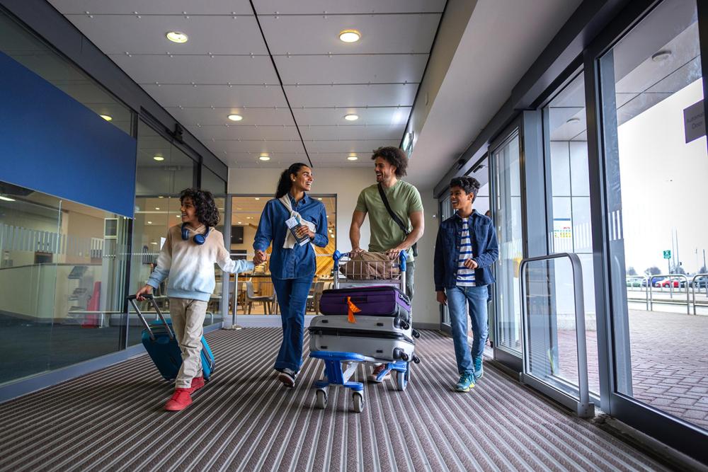 Une famille composée de quatre personnes marche le long d'un couloir d'aéroport, un homme pousse un chariot contenant des valises, tandis qu'une femme tient la main d'un jeune homme en souriant 
