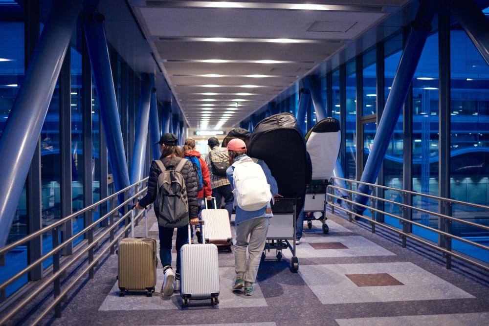 Gruppe von Personen mit Rollkoffern und Kinderwagen geht durch einen beleuchteten, überdachten Gang mit blauen Stahlträgern