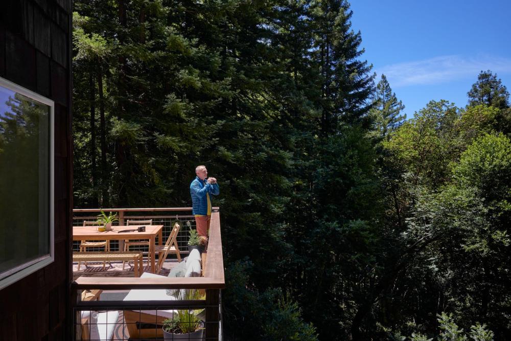 Mann auf Holzterrasse mit Fernglas blickt in dichten Wald mit hohen Nadelbäumen