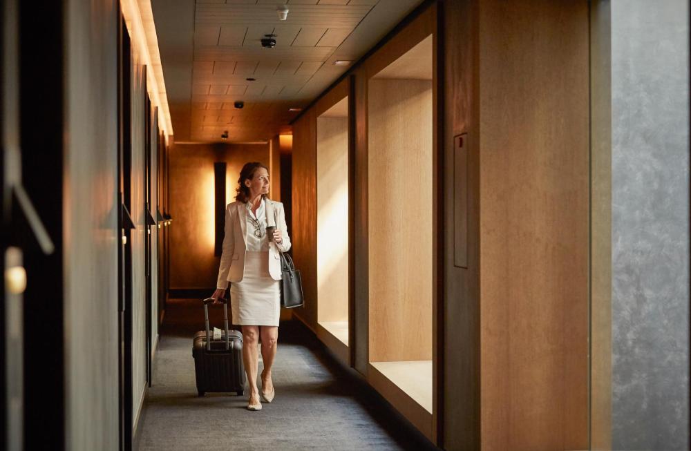 Frau in Businesskleidung zieht einen Rollkoffer und trägt eine Handtasche in einem Hotelflur mit großen Fenstern