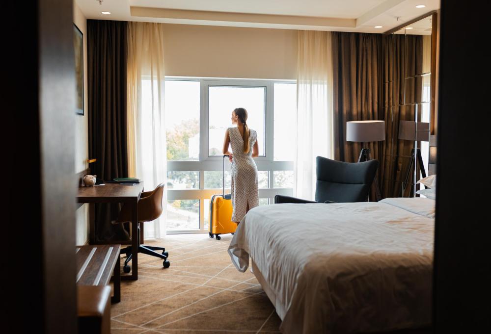 Frau mit langem Zopf und weißem Kleid steht mit gelbem Koffer vor einem großen Fenster in einem Hotelzimmer mit Bett, Schreibtisch und Sessel