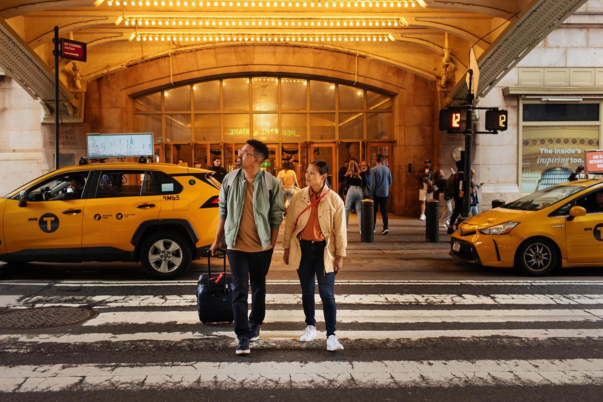 A couple with a suitcase cross a street near a yellow cab